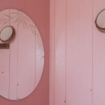 12 out of 15 photographs. Another corner of the pink bathroom is shown, with a small, brass articulating mirror on one side, which is also reflected in a round mirror on the other face of the corner. This mirror is etched with an agricultural motif.